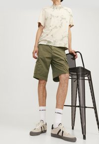 Osoba ve vrstvě z béžového trička s tie-dye, olivovém green cargo šortkách, bílých značkových ponožkách a béžových teniskách, stojící vedle černé kovové vysoké židle.