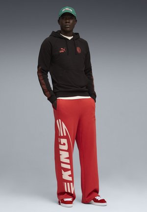 Homme debout portant un sweat à capuche noir AC Milan, un pantalon de survêtement rouge avec le texte "KING", des baskets rouges, et une casquette verte "AC MILAN" sur fond gris.
