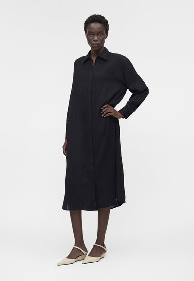 Γυναίκα που στέκεται με το χέρι στη μέση, φοράει ένα μακρύ μαύρο πουκαμισοφόρεμα με κουμπιά και κρεμ αιχμηρές μπαλαρίνες μπροστά σε απλό γκρι φόντο.