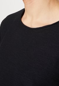 Svart stickad tröja med rund halsringning. Tyget verkar mjukt och texturerat, med ett fint stickat mönster. Nära vy av kragområdet.