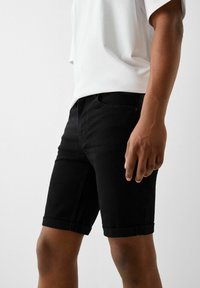 Černé denimové šortky s ohrnutým lemem, které mají klasický pětimístný design a hladkou texturu. Nosené s bílou košilí s krátkým rukávem.