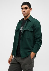 Zielona, teksturowana koszula z guzikami, z dwiema kieszeniami na piersi, noszona na czarnej koszulce z grafiką. Miękka tkanina, kołnierzowy krój oraz dopasowane rękawy.