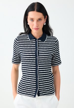 Femme aux longs cheveux foncés portant une chemise à manches courtes rayée bleu marine et blanc boutonnée et un pantalon blanc, debout devant un fond uni.
