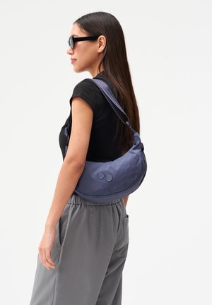 Frau mit schwarzer Sonnenbrille und schwarzem Kurzarmoberteil trägt eine marineblaue, halbmondförmige Umhängetasche mit zwei runden Details.