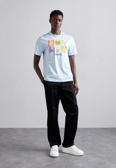 Ljusblå bomulls-T-shirt med ett flerfärgat grafiskt tryck med olika mönster, kombinerad med svarta byxor och vita sneakers.
