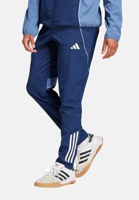 Marineblauwe sportbroek met zijsstripes, gemaakt van een gladde stof; heeft een aangesloten ontwerp en een bijpassend wit Adidas-logo. Witte sneakers met zwarte strepen.