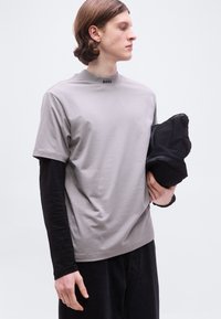 Νέος άνδρας που φορά γκρι κοντομάνικο πουκάμισο πάνω από μαύρο μακρυμάνικο μπλουζάκι, κρατώντας ένα διπλωμένο μαύρο σακάκι, κοιτάζοντας προς τα δεξιά μπροστά σε λευκό φόντο.