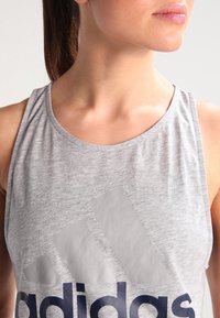 Top atlético sin mangas de color gris, con un escote redondo y el logo prominente de Adidas en azul oscuro en el pecho. Textura de tela suave.