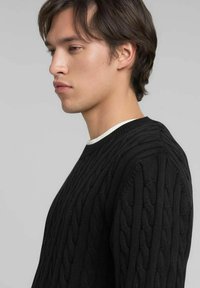 Pull noir en maille avec motif torsadé, col et poignets côtelés. Coupe ajustée vue de profil, mettant en valeur la texture.