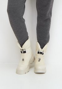 Białe ocieplane buty o puszystym designie, z czarnym akcentem i srebrnymi wykończeniami, zestawione z dopasowanymi szarymi spodniami.