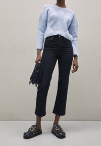 Maglione lavorato a maglia di colore azzurro chiaro con dettagli a costine, jeans neri a vita alta a palazzo e scarpe nere slip-on con hardware dorato. Tiene una borsa con frange.