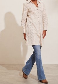 Abito chemisier bianco in pizzo con maniche lunghe e dettaglio dei bottoni, abbinato a jeans chiari a zampa d'elefante e stivaletti con tacco marroni.