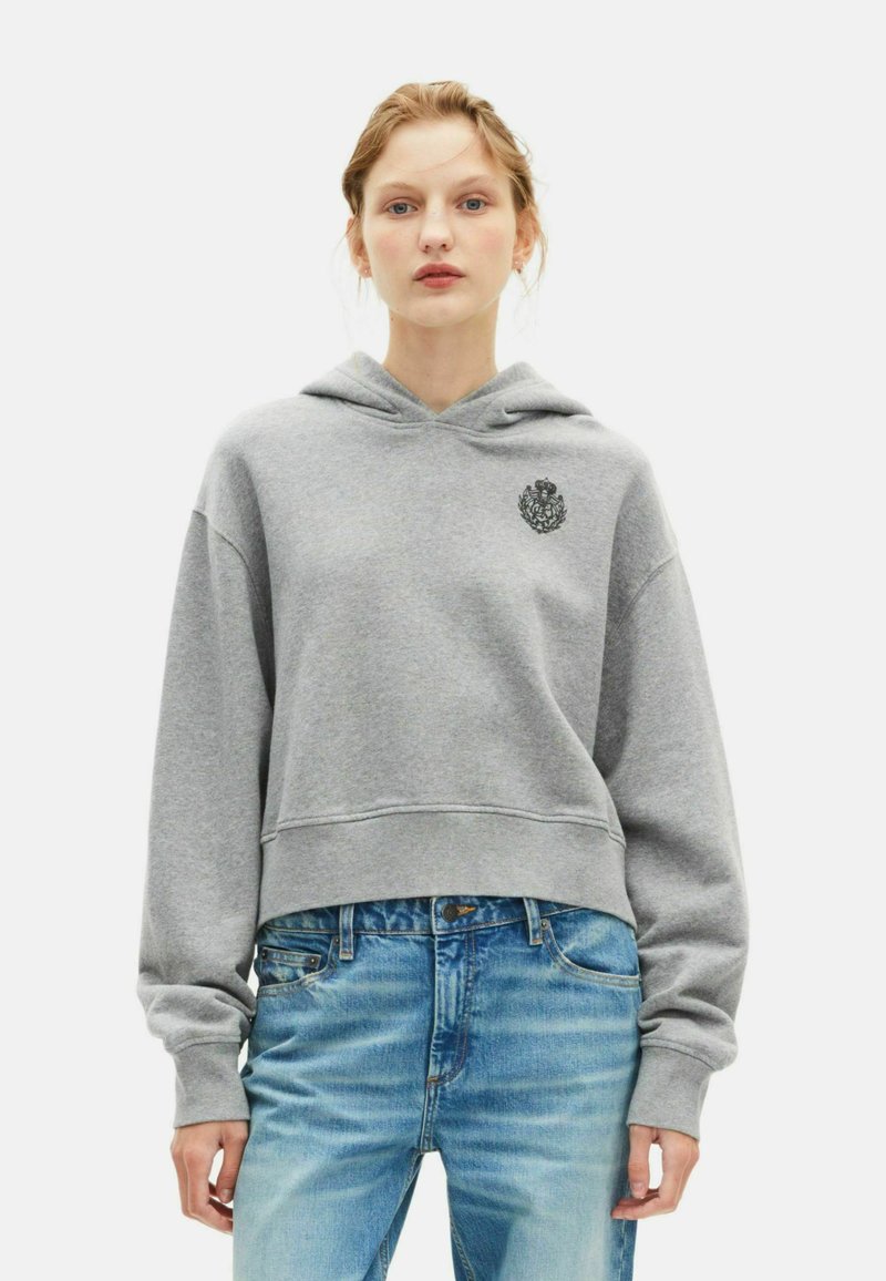 Felpa corta grigia con cappuccio, caratterizzata da polsini a coste e un emblema nero decorativo sul lato sinistro del petto; abbinata a jeans in denim blu chiaro.