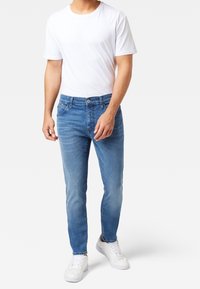 Man som bär ljusblå slim-fit jeans, vit t-shirt och vita sneakers, står mot en enfärgad bakgrund.