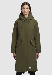 Parka vert olive avec une capuche, poches avant, fermeture à cordon de serrage et détails zippés. Tissu lisse avec un design simple et utilitaire.