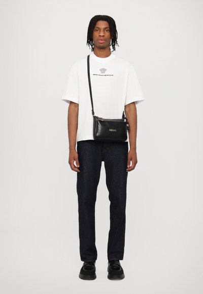 Borsa a tracolla in pelle nera con hardware dorato, forma rettangolare, texture liscia e dettaglio del logo. Indossata con una t-shirt bianca e jeans scuri.