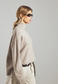 Beige Strickpullover mit hohem Kragen, oversized Passform, gerippten Bündchen, kombiniert mit cremefarbenen Hosen und schwarzen Sonnenbrillen. Glatte Textur.