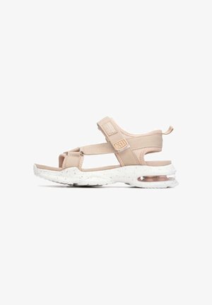 Sandalias deportivas beige con correas ajustables, una parte superior de tela texturizada y una suela exterior blanca moteada que presenta una unidad de aire visible en el talón.