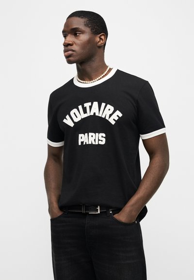 Άνδρας που φοράει μαύρο t-shirt Voltaire Paris με λευκή περιγραφή, μαύρα τζιν, μαύρη ζώνη και περιδέραιο με πέρλες, στέκεται με τα χέρια στις τσέπες.