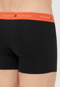 Schwarze Boxer-Shorts mit glatter Textur und einem orangen Bund, auf dem das "TOMMY HILFIGER"-Logo in Weiß prangt.