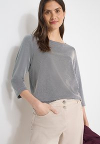 Femme aux longs cheveux noirs portant un haut gris rayé ajusté à manches trois-quarts et un pantalon beige clair, tenant une veste pliée de couleur violet foncé.