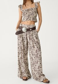 Vrouw in een bijpassende bruine en crèmekleurige paisley-print crop top en wijde broek met een gevlochten riem en houten platform sandalen.