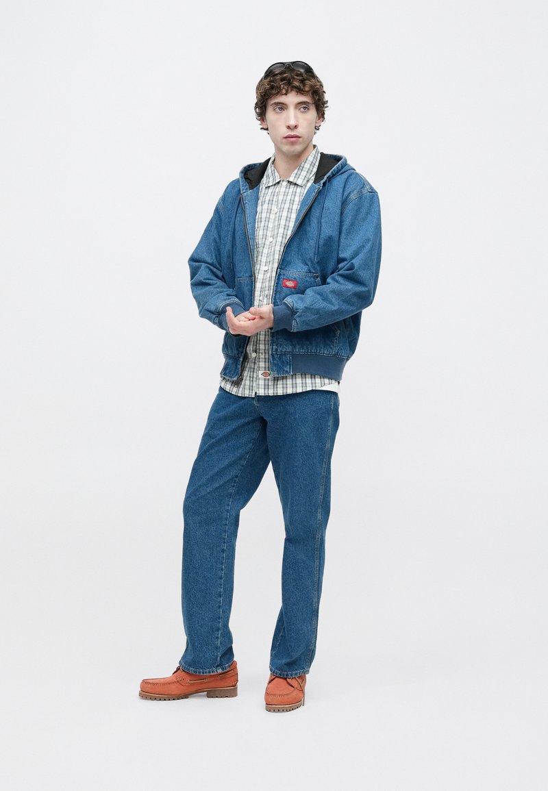 Jeansjacke in mittlerem Blau mit schwarzer Kapuze, darunter kariertes Hemd, blaue Jeans und orange-braune Schuhe. Mit Vordertasche und Reißverschluss.