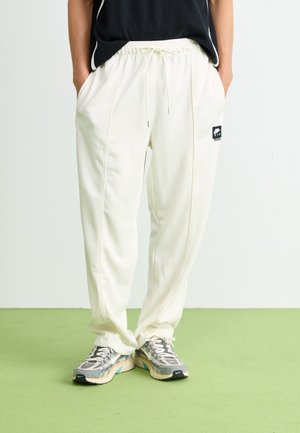 Person wearing cream-colored Nike Air sweatpants and gray sneakers standing on a green floor with hands in pockets.