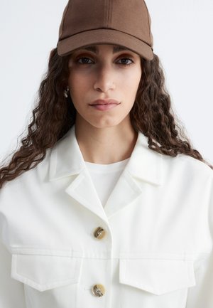 Femme aux longs cheveux bouclés portant une casquette marron, une veste blanche boutonnée et une chemise blanche, face à l’avant sur un fond clair uni.