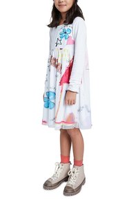 Biała sukienka z kolorowymi wzorami, w tym kwiatami i gwiazdami; długie rękawy; połączona z beżowymi butami i różowymi skarpetkami.