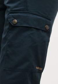 Dark blue fabric cargo pocket on pants with two metal button closures and orange "G-1000" text embroidered near bottom edge.