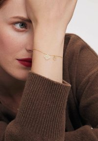 Złota bransoletka z kwiatowym wzorem z dwoma zawieszkami, jedną większą i jedną mniejszą, noszona na lekko teksturowanym odcieniu skóry.