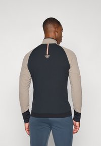 Mustikas ja beež pikkade varrukatega spordijakk, millel on kõrge kaelus, tekstuuriga kangas ja roosa esiletõstmisriie seljal, featuring a logo.
