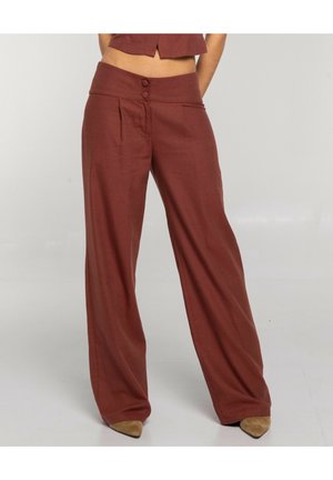 Mujer con pantalones marrones de talle alto y pierna ancha con dos botones en la cintura, combinados con zapatos color canela de punta afilada sobre un fondo liso.