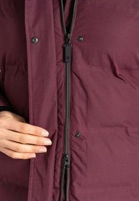 Bordeauxfarbene isolierte Jacke mit Reißverschluss, Druckknöpfen und einer weichen Textur. Die Jacke hat ein schlankes Design und eine figurbetonte Silhouette.