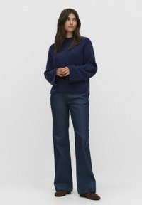 Granatowy sweter wełniany z szerokimi rękawami, zestawiony z ciemnoniebieskimi dzwonami. Model stoi na jasnym tle.