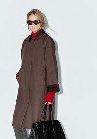 Cappotto marrone con colletto, tasche laterali e polsini scuri a contrasto, abbinato a un maglione rosso e a una borsa a tracolla nera con texture a coccodrillo.