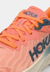 Hoka running shoe in coral and orange, featuring textured mesh upper, thick sole, and contrasting blue branding. Laces are bright orange.