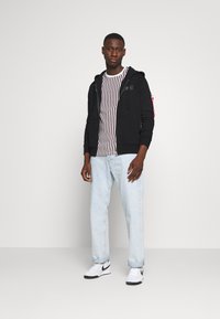 Black zip-up hoodie with front pockets over a white striped t-shirt, paired with light blue jeans and white sneakers with black accents.