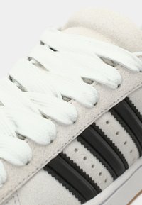 Λευκό σουέτ αθλητικό παπούτσι με μαύρες δερμάτινες ρίγες, επίπεδες λευκές κορδόνια, διακοσμητικές τρύπες και λαστιχένια σόλα. Κοντινή άποψη του σχεδιασμού του πάνω μέρους.