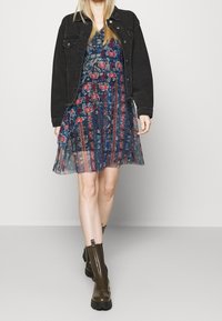 Kvinna klädd i en svart jeanjacka över en blå mönstrad klänning med röda och beige mönster, ihop med mörkbruna ankelboots.