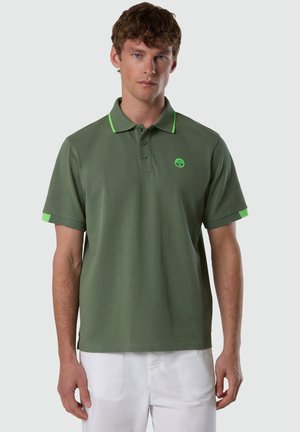 Giovane uomo che indossa una polo verde oliva con dettagli verde neon e pantaloni bianchi, in piedi contro uno sfondo chiaro e uniforme.