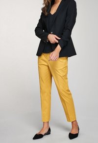 Blazer noir ajusté sur un top sans manches, associé à un pantalon jaune slim et des ballerines noires. Tissu lisse avec des lignes épurées.