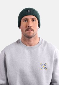 Sweatshirt gris avec un motif floral sur la poitrine gauche ; bonnet vert foncé avec un patch logo ; fond uni.