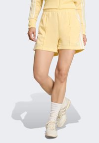 Gele sportshorts met een gladde textuur, voorzien van witte biesstrepen aan de zijkanten en een elastische tailleband. Gecombineerd met lichtgekleurde sneakers en sokken.