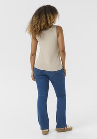 Beige mouwloze top, blauwe flared jeans met een nauwsluitende taille en textuur breisel. Lichtbruine schoenen met rode en witte accenten.