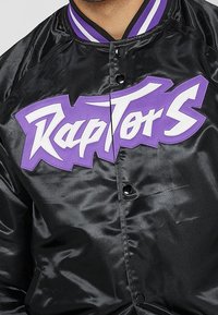 Chaqueta de satén negra con el logotipo "Raptors" bordado en púrpura, botones de presión y un cuello acanalado con rayas púrpuras y blancas.