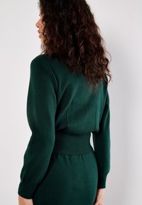 Kobieta z kręconymi włosami, ubrana w ciemnozieloną, długą dzianinową sukienkę z dopasowaną talią, zwrócona w lewo na tle jasnym.