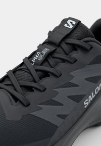 Black athletic shoe featuring a textured upper, mesh panels, flat laces, and a distinctive jagged design pattern. "SALOMON" branding is visible.
