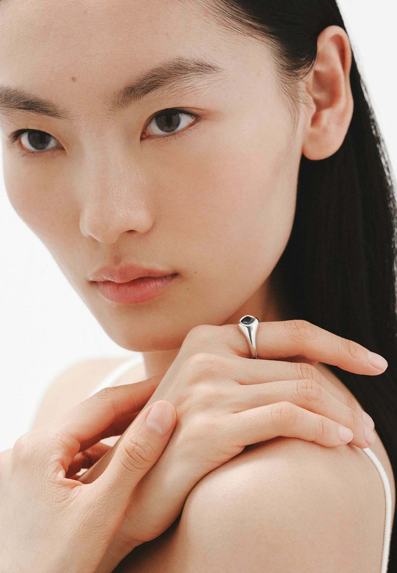 Anillo de plata con un engaste de piedra negra, mostrado en una mano descansando sobre la piel. Diseño suave y elegante; estilo minimalista; sin patrones intrincados.